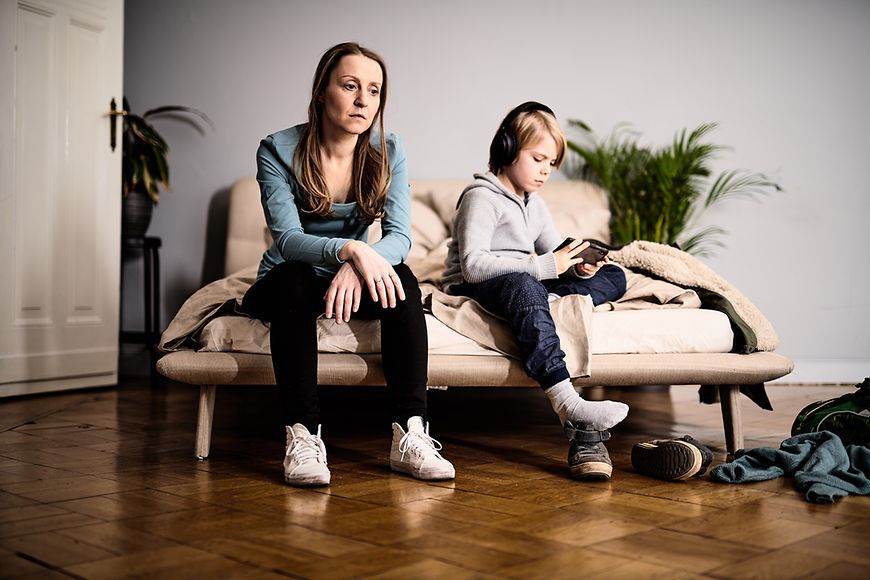 Ein Mutter schaut auf einer Couch ins Leere, neben ihr sitzt ein Junge, der ein Computerspiel spielt