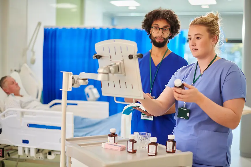 Zwei pflegende Personen und ein Mensch in einem Krankenbett im Hintergrund