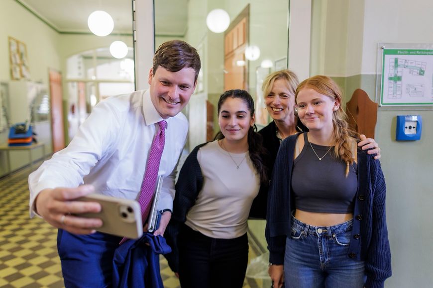 Karin Prien und Conrad Clemens stehen in einem Klassenzimmer und unterhalten sich mit Schülerinnen und Schülern