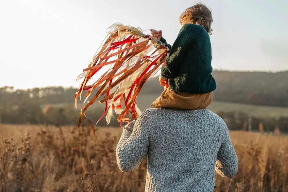 Ein Kind sitzt auf den Schultern eines Erwachsenen. Das Kind hält einen gebastelten Stock mit bunten Bändern in der Hand.