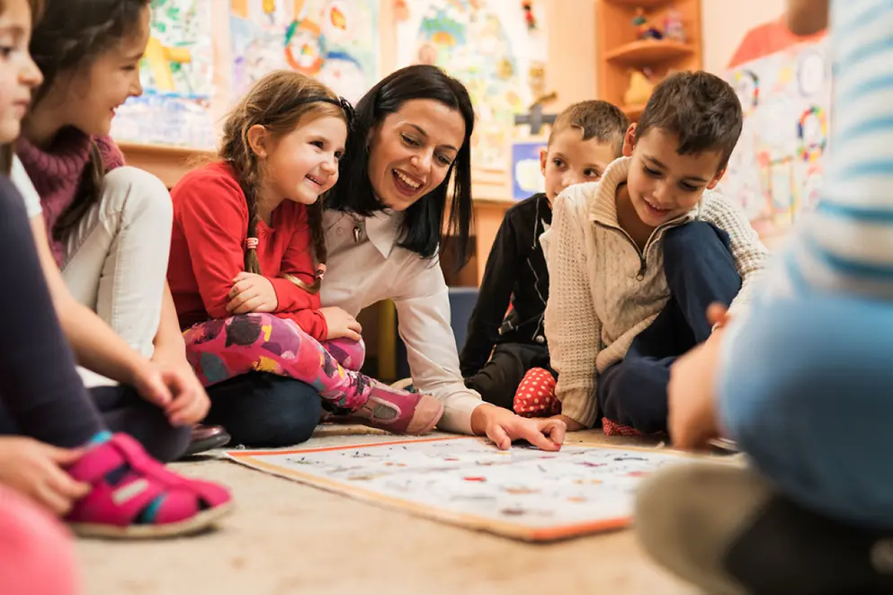 Eine Frau sitzt mit lachenden Kindern auf dem Boden in einer Kindertageseinrichtung.