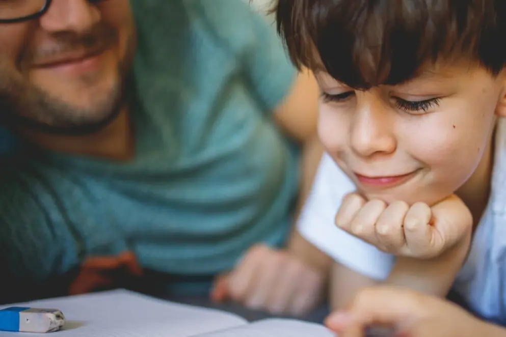 Ein Kind schaut lächelnd in ein Buch und stützt den Kopf auf die Hand. Der Vater lächelt.