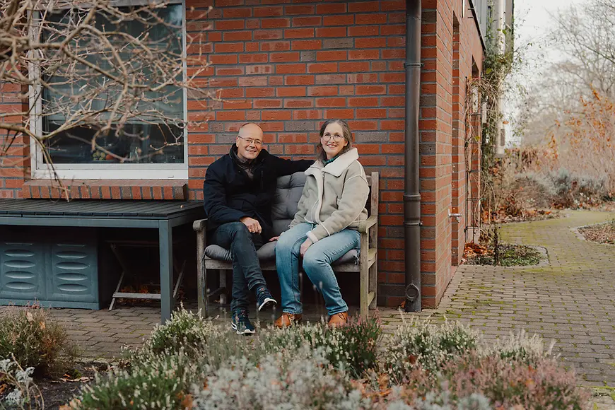 Ein älteres Paar sitzt auf einer Bank in einem Garten und lächelt in die Kamera