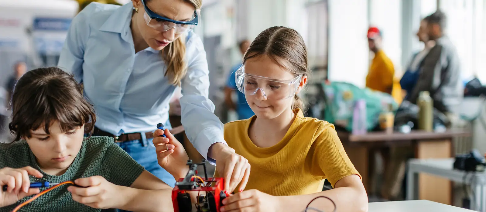 Eine Erwachsene mit Schutzbrille hilft zwei Kindern beim Zusammenbau von elektronischen Teilen.