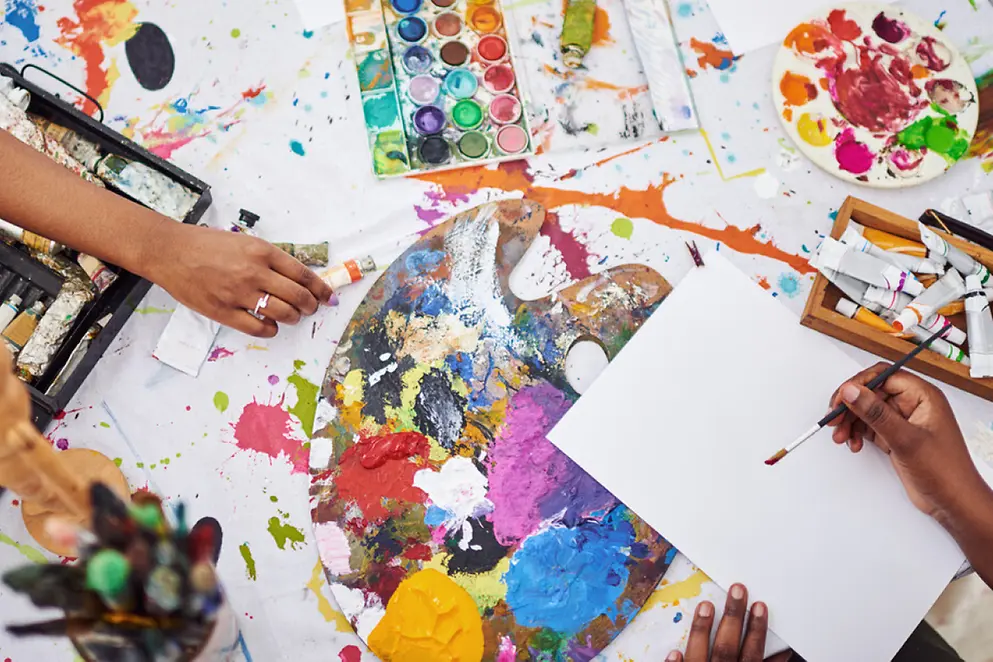 Eine Hand beim kreativen Arbeiten mit bunten Farben, Papier und Pinsel