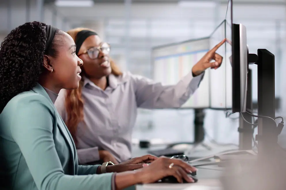 Eine Jugendliche und eine Frau sitzen vor einem Monitor. Die Frau erklärt etwas.