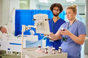 Zwei pflegende Personen und ein Mensch in einem Krankenbett im Hintergrund